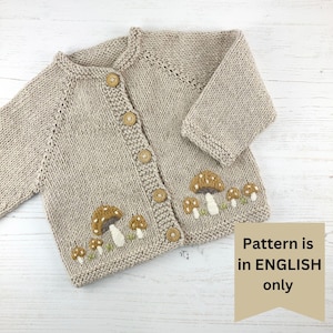 May include: A beige knitted baby cardigan with a row of embroidered mushrooms on the bottom. The cardigan has wooden buttons down the front.
