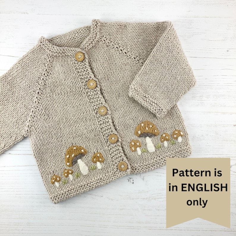 May include: A beige knitted baby cardigan with a row of embroidered mushrooms on the bottom. The cardigan has wooden buttons down the front.