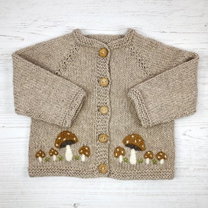 May include: A beige knitted cardigan with a mushroom design. The button-up sweater features brown and white embroidered mushrooms with green grass accents. The cardigan has long sleeves and wooden buttons.