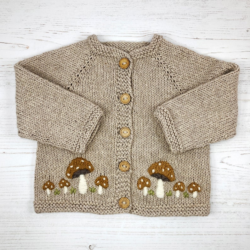 May include: A beige knitted cardigan with a mushroom design. The button-up sweater features brown and white embroidered mushrooms with green grass accents. The cardigan has long sleeves and wooden buttons.