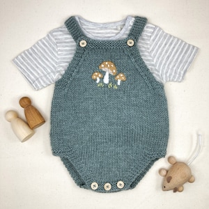 May include: A teal knitted baby romper with embroidered mushrooms, paired with a striped short-sleeve shirt. The romper has wooden button closures and is accompanied by wooden toys, including a mouse and two peg dolls.