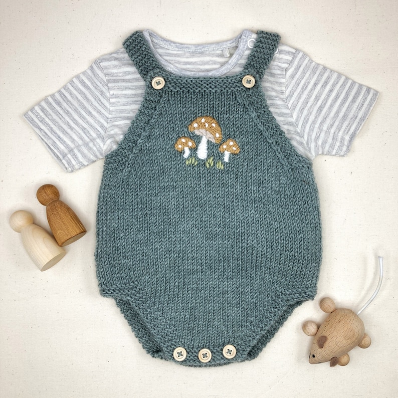 May include: A teal knitted baby romper with embroidered mushrooms, paired with a striped short-sleeve shirt. The romper has wooden button closures and is accompanied by wooden toys, including a mouse and two peg dolls.