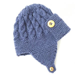 May include: A blue knitted hat with a cable knit pattern and two wooden buttons. The hat has a long flap that can be tied under the chin.