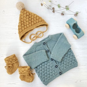 Può includere: Un set di vestiti per neonati in maglia di colore blu chiaro e giallo senape. Il set comprende un cardigan, un cappello e scarpine.