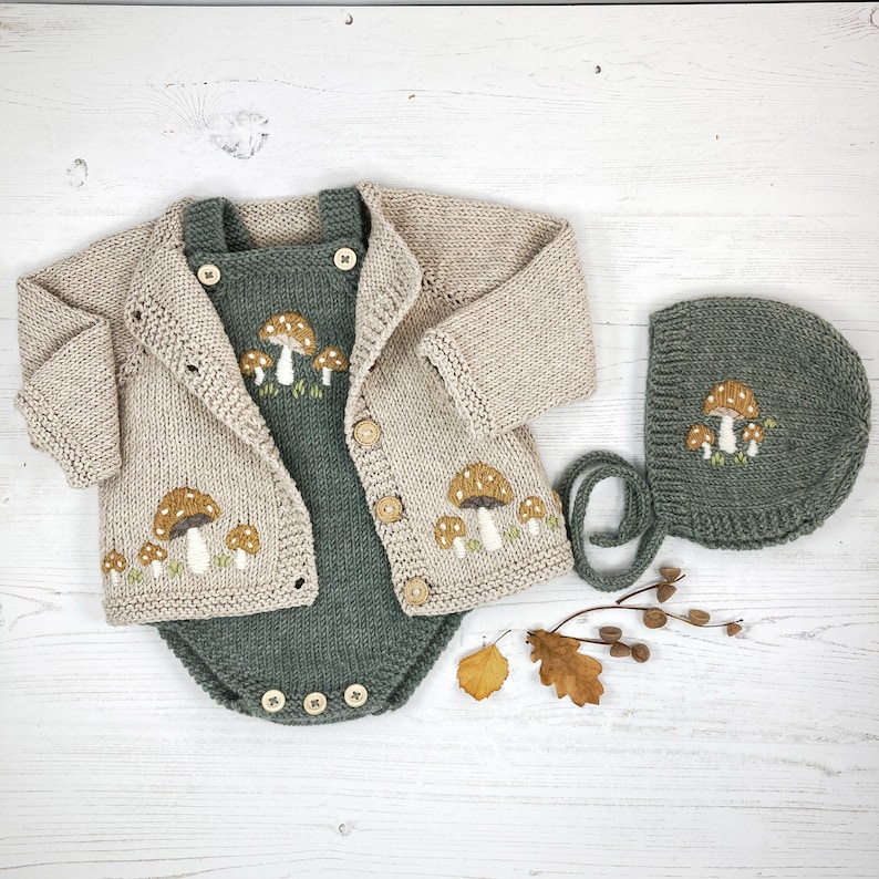 May include: A three-piece baby outfit set featuring a beige cardigan, a green romper, and a matching bonnet. The set is knit and embroidered with mushroom designs. The romper has wooden buttons.