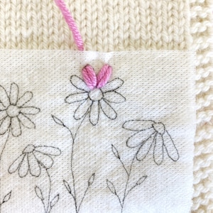May include: A close-up of a white fabric with black embroidered daisies and a pink embroidered daisy with a pink thread.