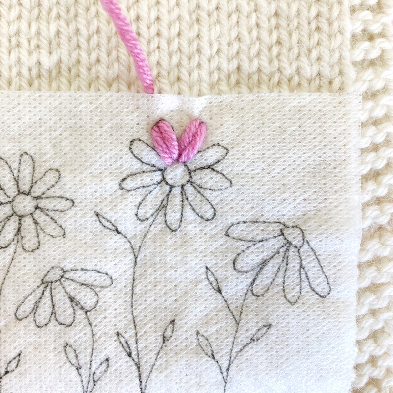 May include: A close-up of a white fabric with black embroidered daisies and a pink embroidered daisy with a pink thread.
