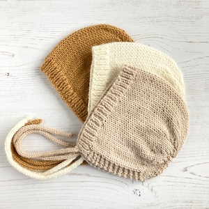 Op de afbeelding: Drie gebreide baby mutsjes in beige, crème en mosterdgeel met strikjes. De mutsjes zijn op elkaar gestapeld.