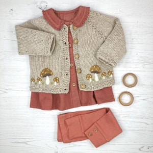 May include: A beige knitted cardigan with mushroom embroidery, a coral colored dress with a ruffled collar, and coral colored leggings.