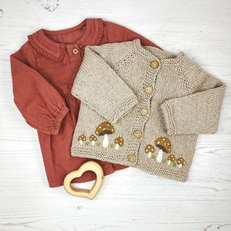 May include: A beige knitted cardigan with mushroom embroidery and brown buttons. The cardigan is paired with a red corduroy shirt with a ruffled collar.