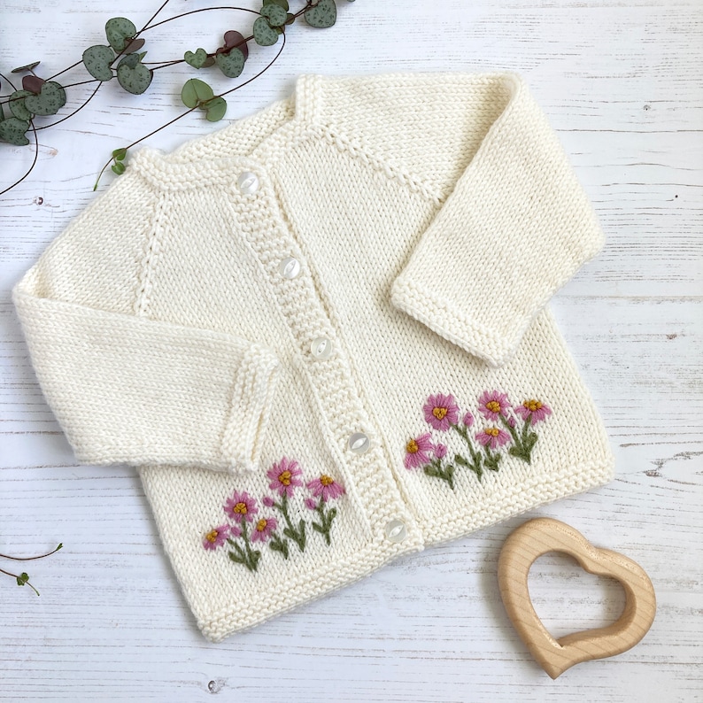 May include: A white knitted cardigan with a button closure and embroidered pink flowers.