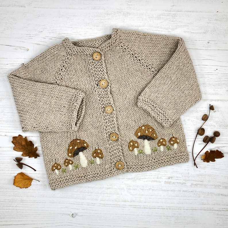 May include: A beige knitted cardigan with embroidered mushrooms in brown and green. The cardigan has wooden buttons.