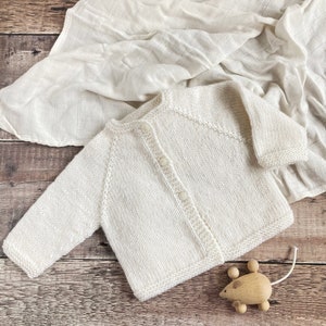 May include: A cream-colored, long-sleeved knit cardigan featuring a round neckline and a front button closure with three matching buttons. The garment is crafted with a textured knit pattern and ribbed edges along the cuffs and hemline.