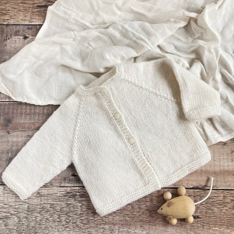 May include: A cream-colored, long-sleeved knit cardigan featuring a round neckline and a front button closure with three matching buttons. The garment is crafted with a textured knit pattern and ribbed edges along the cuffs and hemline.