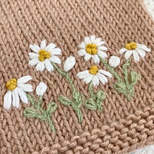 May include: A close-up of a brown knitted fabric with embroidered white daisies. The daisies have yellow centers and green stems.