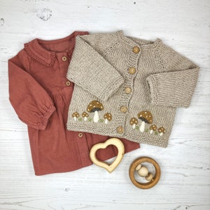 May include: A beige knitted cardigan with mushroom embroidery and a red button-down shirt with a ruffled collar.  The cardigan has a button closure and the shirt has a button closure.  There are two wooden toys in the image, a heart-shaped teether and a wooden rattle.