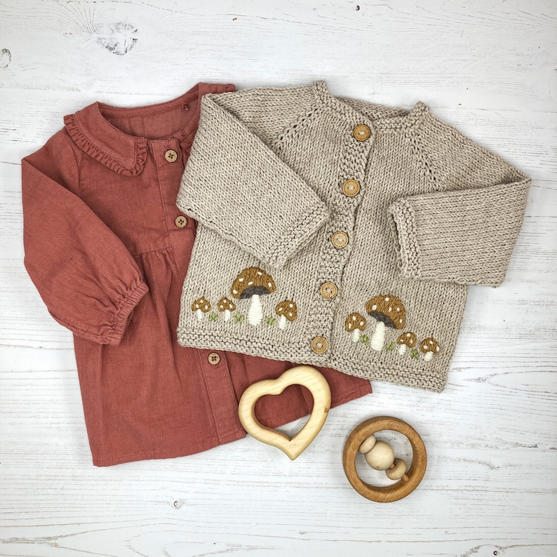 May include: A beige knitted cardigan with mushroom embroidery and a red button-down shirt with a ruffled collar.  The cardigan has a button closure and the shirt has a button closure.  There are two wooden toys in the image, a heart-shaped teether and a wooden rattle.
