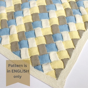 K&ouml;nnte beinhalten: Eine gestrickte Babydecke mit einem gewebten Muster in den Farben Gelb, Blau, Grau und Wei&szlig;. Die Decke hat einen wei&szlig;en Rand.