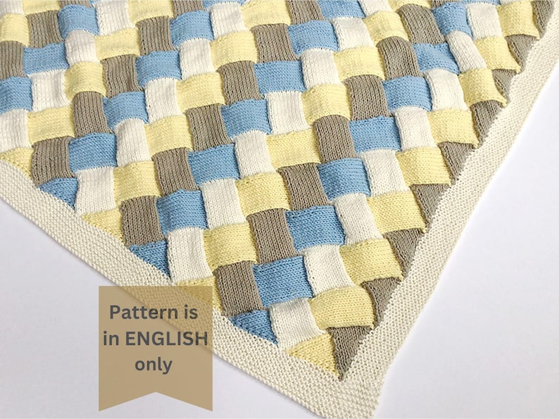 K&ouml;nnte beinhalten: Eine gestrickte Babydecke mit einem gewebten Muster in den Farben Gelb, Blau, Grau und Wei&szlig;. Die Decke hat einen wei&szlig;en Rand.