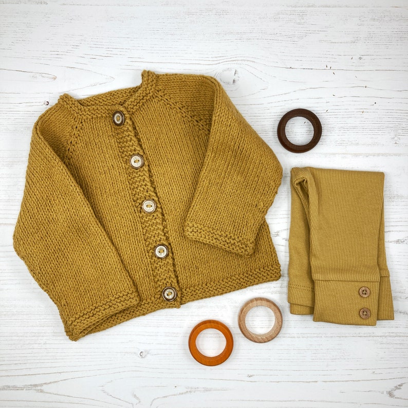 May include: A mustard yellow knitted cardigan with five light-colored buttons, a pair of matching mustard yellow ribbed leggings with two small buttons at the cuffs, and three wooden rings in dark brown, light brown, and orange.