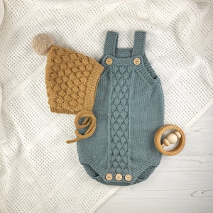 Può includere: Un set di abbigliamento per neonati in maglia blu e marrone. Il set comprende un body in maglia blu con bottoni e un cappello in maglia marrone con un pon pon.