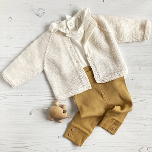 May include: A cream-colored knit cardigan with long sleeves and buttons, layered over a white shirt with a ruffled collar. The outfit includes mustard yellow ribbed leggings with button details at the cuffs and a small wooden mouse toy.