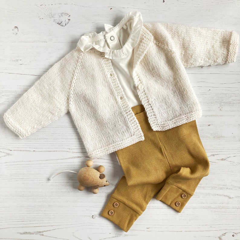 May include: A cream-colored knit cardigan with long sleeves and buttons, layered over a white shirt with a ruffled collar. The outfit includes mustard yellow ribbed leggings with button details at the cuffs and a small wooden mouse toy.