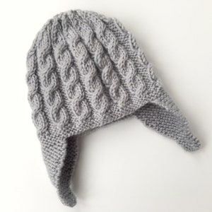 May include: Gray knitted hat with ear flaps. The hat has a cable knit pattern.