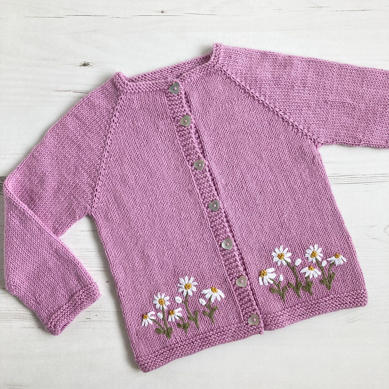 May include: A light purple knitted cardigan with a button closure and embroidered white daisies on the bottom.
