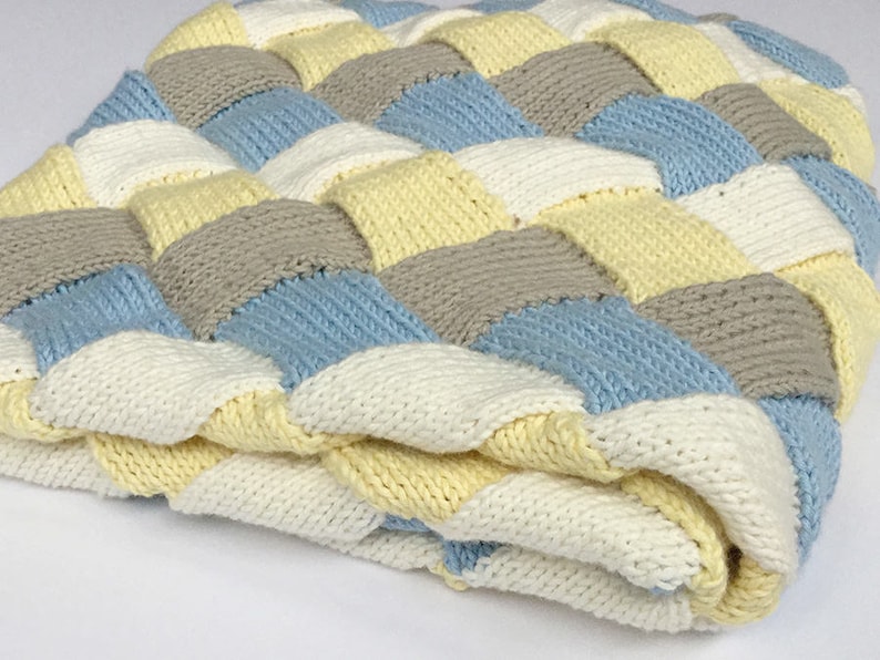 K&ouml;nnte beinhalten: Eine gestrickte Babydecke mit einem gewebten Muster in den Farben Wei&szlig;, Gelb, Hellblau und Grau.
