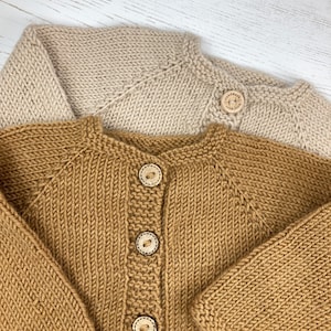 May include: Two knitted cardigans, one in beige and one in mustard yellow, featuring raglan sleeves and a round neckline. The mustard cardigan has three wooden buttons, while the beige one has a single button engraved with "handmade with love".