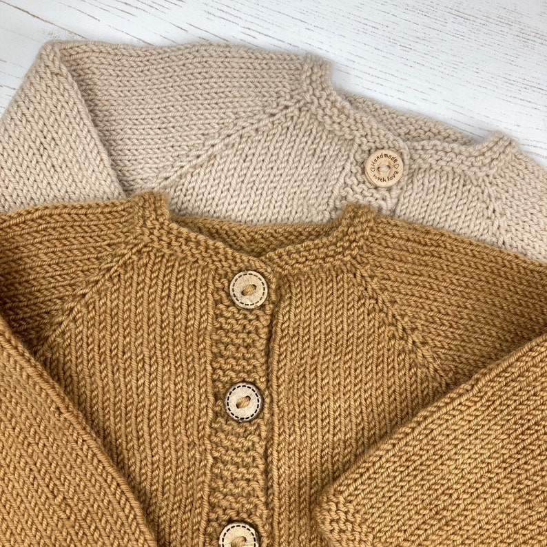 May include: Two knitted cardigans, one in beige and one in mustard yellow, featuring raglan sleeves and a round neckline. The mustard cardigan has three wooden buttons, while the beige one has a single button engraved with "handmade with love".