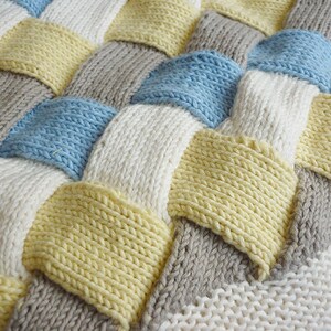 K&ouml;nnte beinhalten: Eine gestrickte Decke mit einem gewebten Muster in den Farben Wei&szlig;, Hellblau, Gelb und Grau.