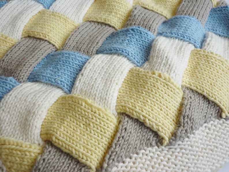 K&ouml;nnte beinhalten: Eine gestrickte Decke mit einem gewebten Muster in den Farben Wei&szlig;, Hellblau, Gelb und Grau.