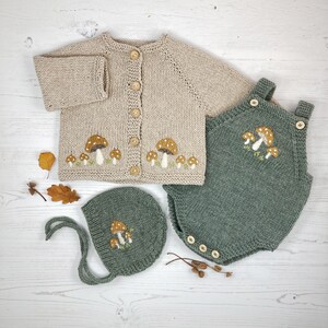 May include: A beige knitted baby cardigan, green romper, and bonnet set. The cardigan and bonnet have embroidered mushroom designs. The romper has a mushroom design on the chest. The set has wooden buttons and is made of soft yarn.