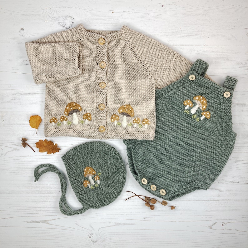 May include: A beige knitted baby cardigan, green romper, and bonnet set. The cardigan and bonnet have embroidered mushroom designs. The romper has a mushroom design on the chest. The set has wooden buttons and is made of soft yarn.