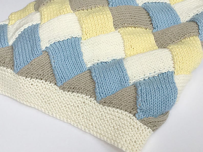 K&ouml;nnte beinhalten: Eine gestrickte Babydecke mit Patchwork-Muster in den Farben Blau, Gelb, Wei&szlig; und Grau.