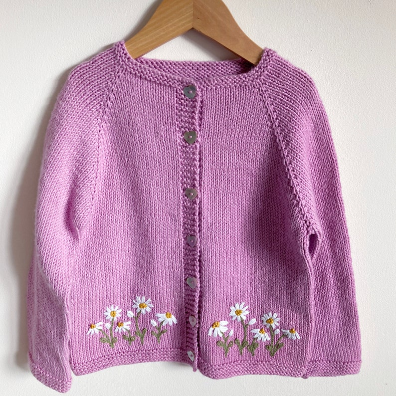 May include: A lavender knitted cardigan with a button closure and embroidered daisies on the bottom hem.