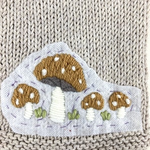 May include: A beige knitted fabric with a patch featuring embroidered mushrooms. The mushrooms have brown caps with white spots, white stems, and green grass. The patch is stitched with purple thread.