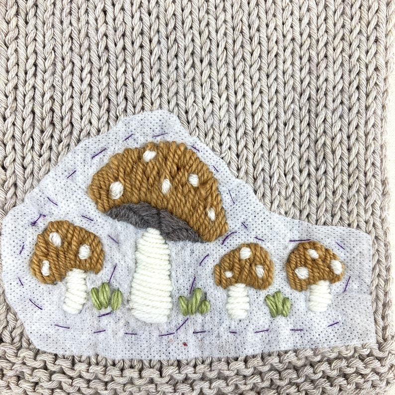 May include: A beige knitted fabric with a patch featuring embroidered mushrooms. The mushrooms have brown caps with white spots, white stems, and green grass. The patch is stitched with purple thread.