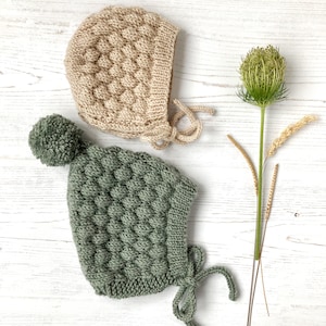 Op de afbeelding: Twee gebreide baby mutsen, één beige en één groen, met een pompon op de groene muts. De mutsen liggen op een wit houten oppervlak.