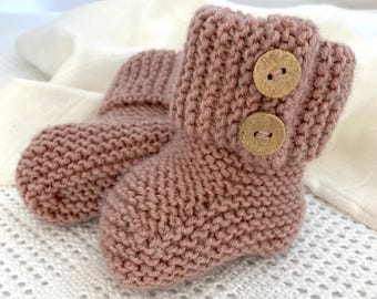 May include: A pair of dusty rose knitted booties featuring a garter stitch texture and a folded cuff. Each bootie is adorned with two round, light brown wooden buttons positioned vertically on the side of the cuff, providing a secure closure.