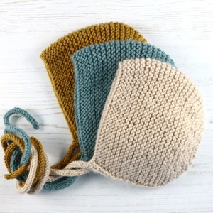 May include: Three knitted baby bonnets in beige, teal, and mustard yellow. The bonnets are made with a simple stitch and have a pointed crown.