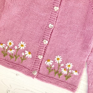May include: A pink knitted cardigan with white embroidered daisies and heart-shaped buttons.