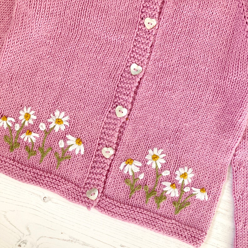 May include: A pink knitted cardigan with white embroidered daisies and heart-shaped buttons.