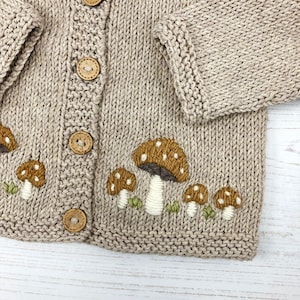 May include: A beige knitted cardigan with embroidered mushrooms in shades of brown, white, and green. The cardigan has wooden buttons.