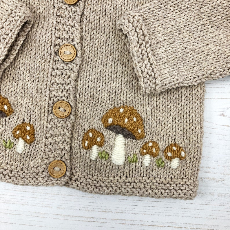 May include: A beige knitted cardigan with embroidered mushrooms in shades of brown, white, and green. The cardigan has wooden buttons.