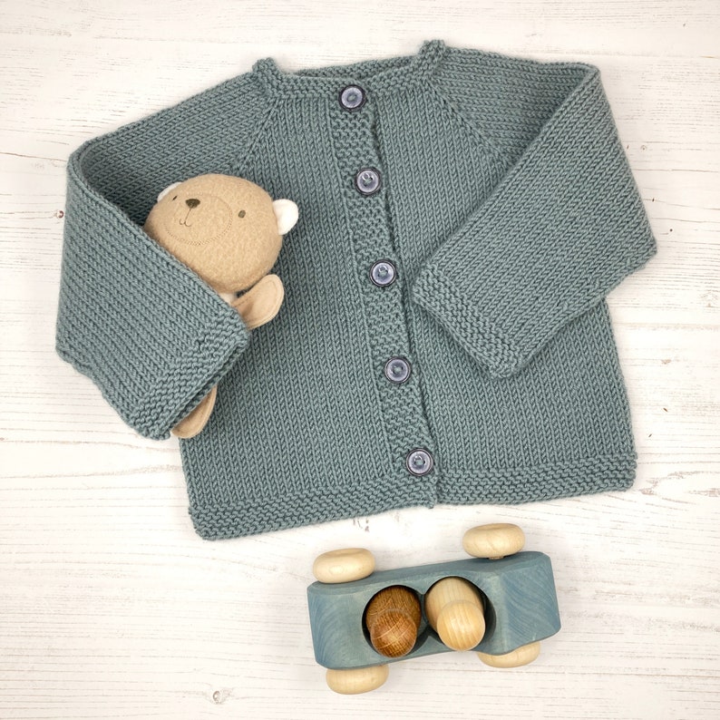 May include: A dusty blue knitted cardigan with long sleeves, a round neckline, and five matching buttons down the front. A small tan plush bear is tucked into the left sleeve, and a blue wooden toy car with two wooden figures sits below.