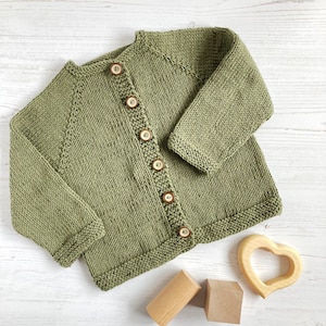 May include: A sage green knitted cardigan featuring long sleeves, a round neckline, and a front button closure with six round brown buttons. The garment is crafted with a textured knit pattern and ribbed edges along the cuffs and hemline.