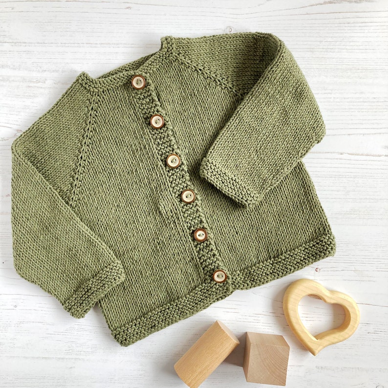 May include: A sage green knitted cardigan featuring long sleeves, a round neckline, and a front button closure with six round brown buttons. The garment is crafted with a textured knit pattern and ribbed edges along the cuffs and hemline.